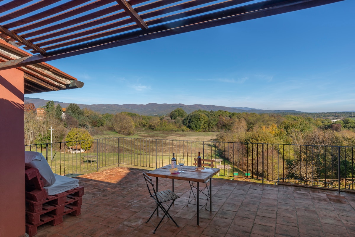A spacious terrace is highlighted, offering a table with chairs set against a scenic view of rolling hills and greenery. Sunlight pours over the area, while a canopy of rustic beams provides shade. The serene landscape invites relaxation and outdoor dining.