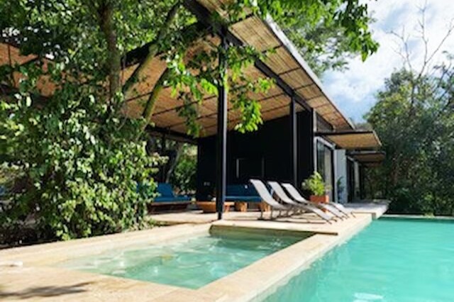 An open area presents a clear view of a refreshing pool and a jacuzzi, surrounded by lush greenery. Comfortable lounge chairs are positioned for relaxation under a covered terrace, allowing for both shade and light from the sky above.