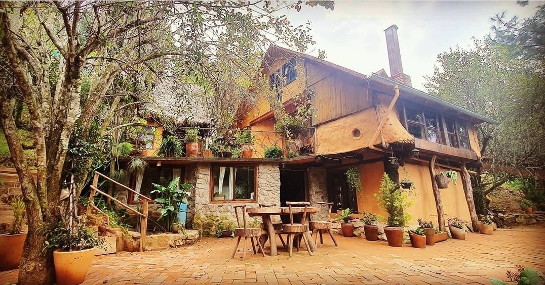 A charming multi-level cabin is surrounded by lush greenery and decorative potted plants. The structure features natural materials, with wooden and stone elements visible. A rustic wooden table with chairs is positioned on a brick patio, inviting outdoor gatherings.