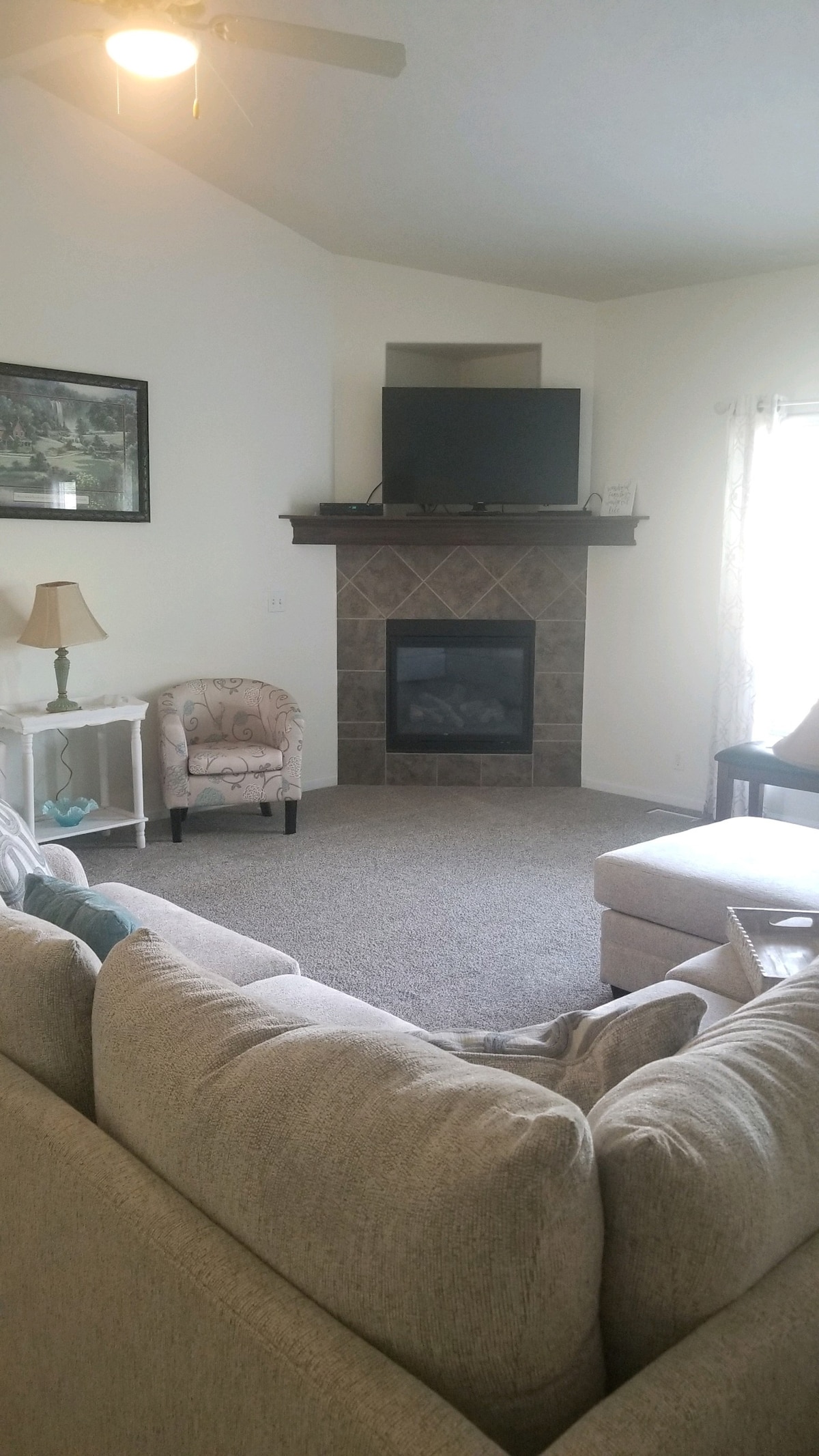 A cozy living room showcases a stone fireplace, with a television mounted above. Soft seating includes a light-colored sofa, a patterned armchair, and a small side table with a lamp. Natural light enters through sheer curtains, creating a bright and inviting ambiance.