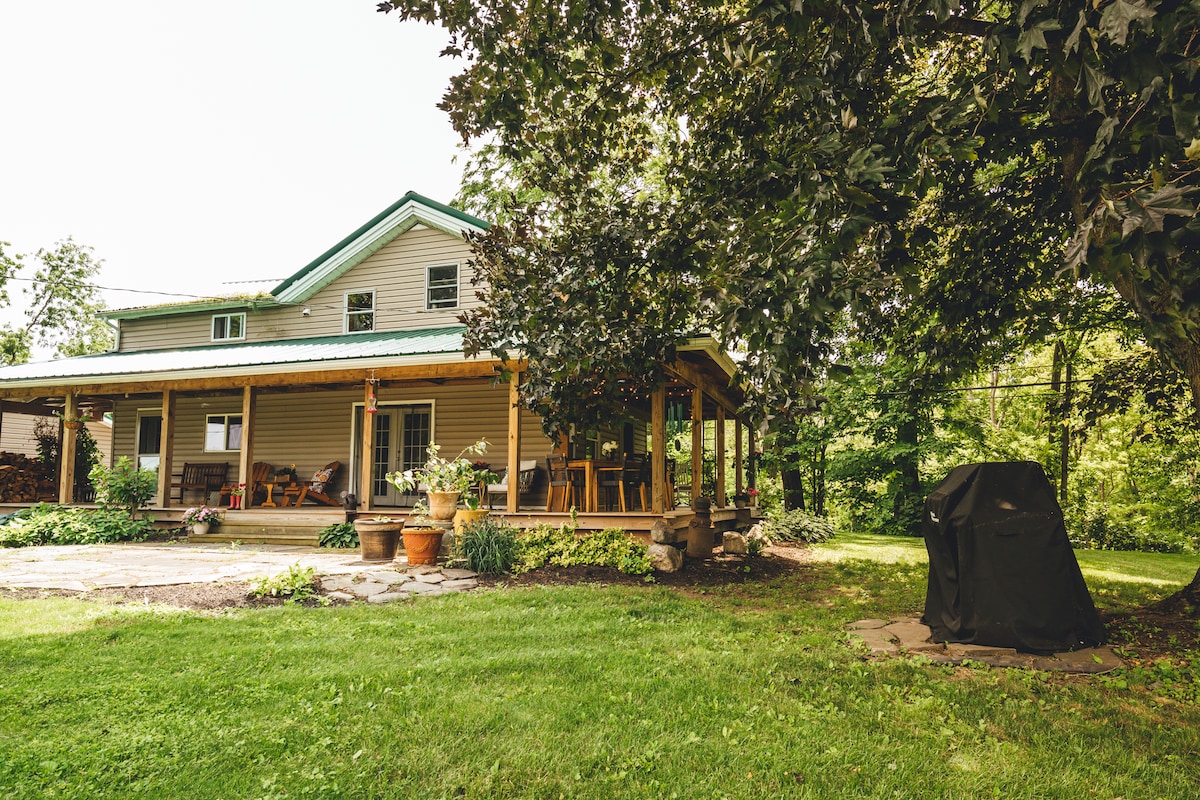 A spacious farmhouse is surrounded by greenery, featuring a welcoming porch with seating and a dining area. Potted plants decorate the outdoor space, while a covered grill is seen on the patio. The well-maintained lawn offers ample room for relaxation.