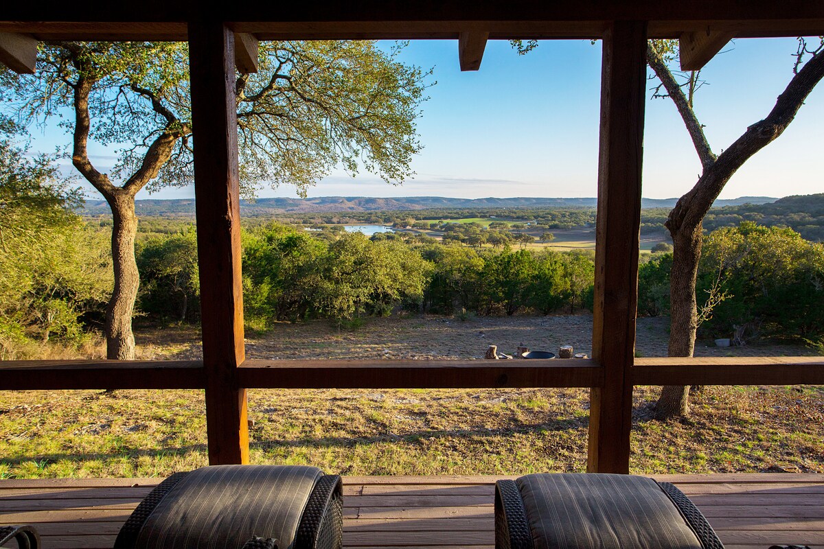 A serene view from a wooden porch reveals rolling hills and lush greenery under an expansive blue sky. The landscape includes a distant water body, framed by trees that enhance the natural beauty of the area.