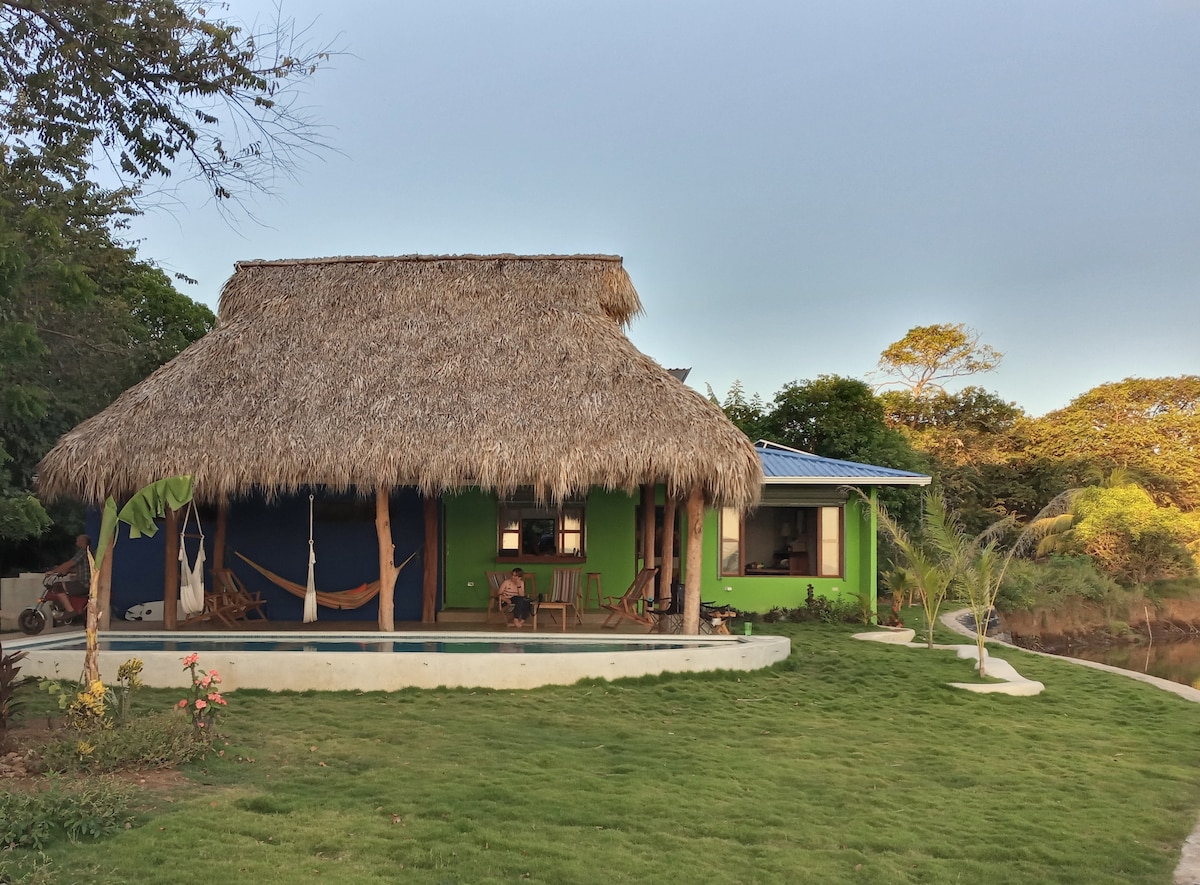 A tropical-inspired structure features a thatched roof and colorful exterior, nestled among lush greenery. A small pool reflects the sky, while several hammocks are arranged on the porch. Walkways meander through the neatly manicured lawn, leading to the surrounding landscape.