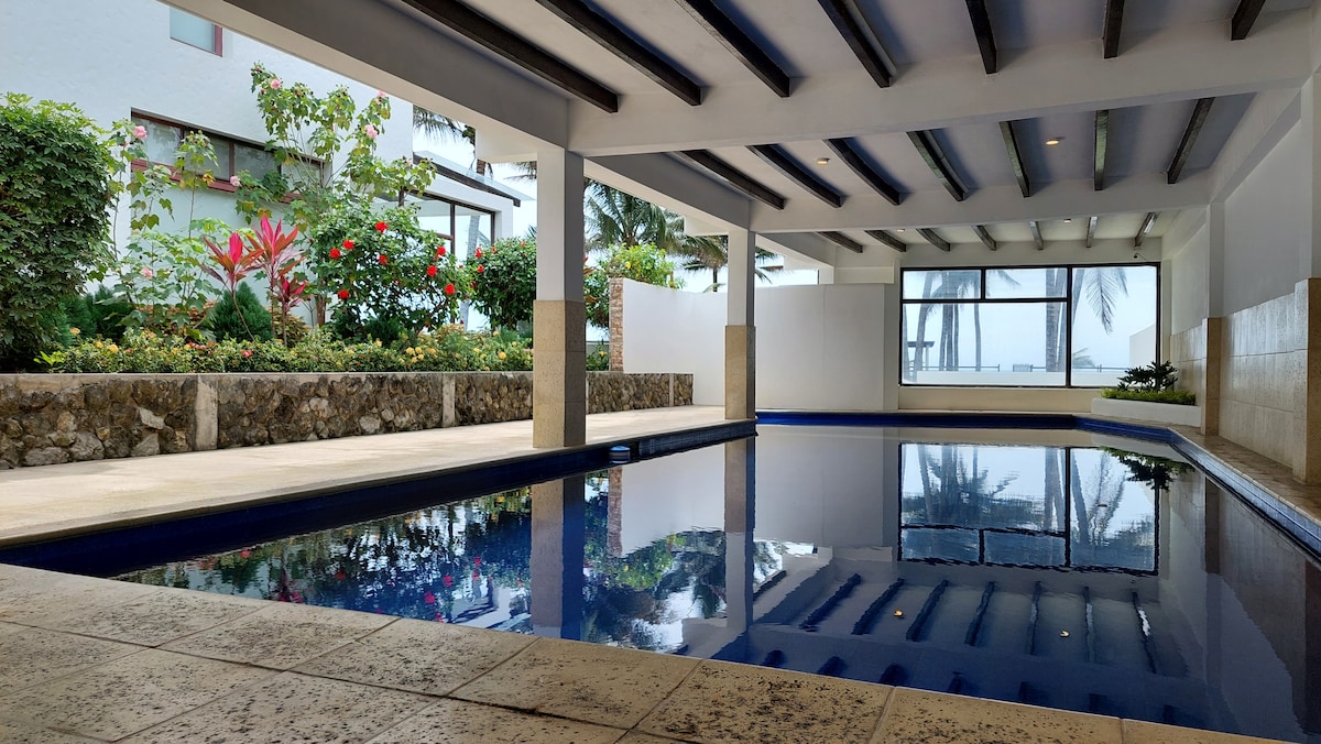 An indoor heated pool is surrounded by spacious seating and lush greenery. Large windows provide natural light and views of palm trees outside, creating a calm and relaxing environment. The water's surface reflects the surrounding area, enhancing the tranquil atmosphere.