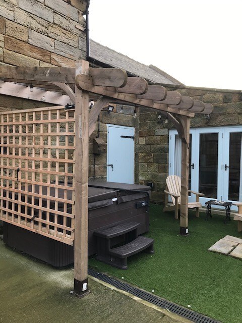 A private hot tub is featured under a wooden pergola, surrounded by timber lattice for privacy. Adjacent to the hot tub, two chairs are positioned on modern artificial grass, creating a relaxing outdoor space suitable for enjoyment.