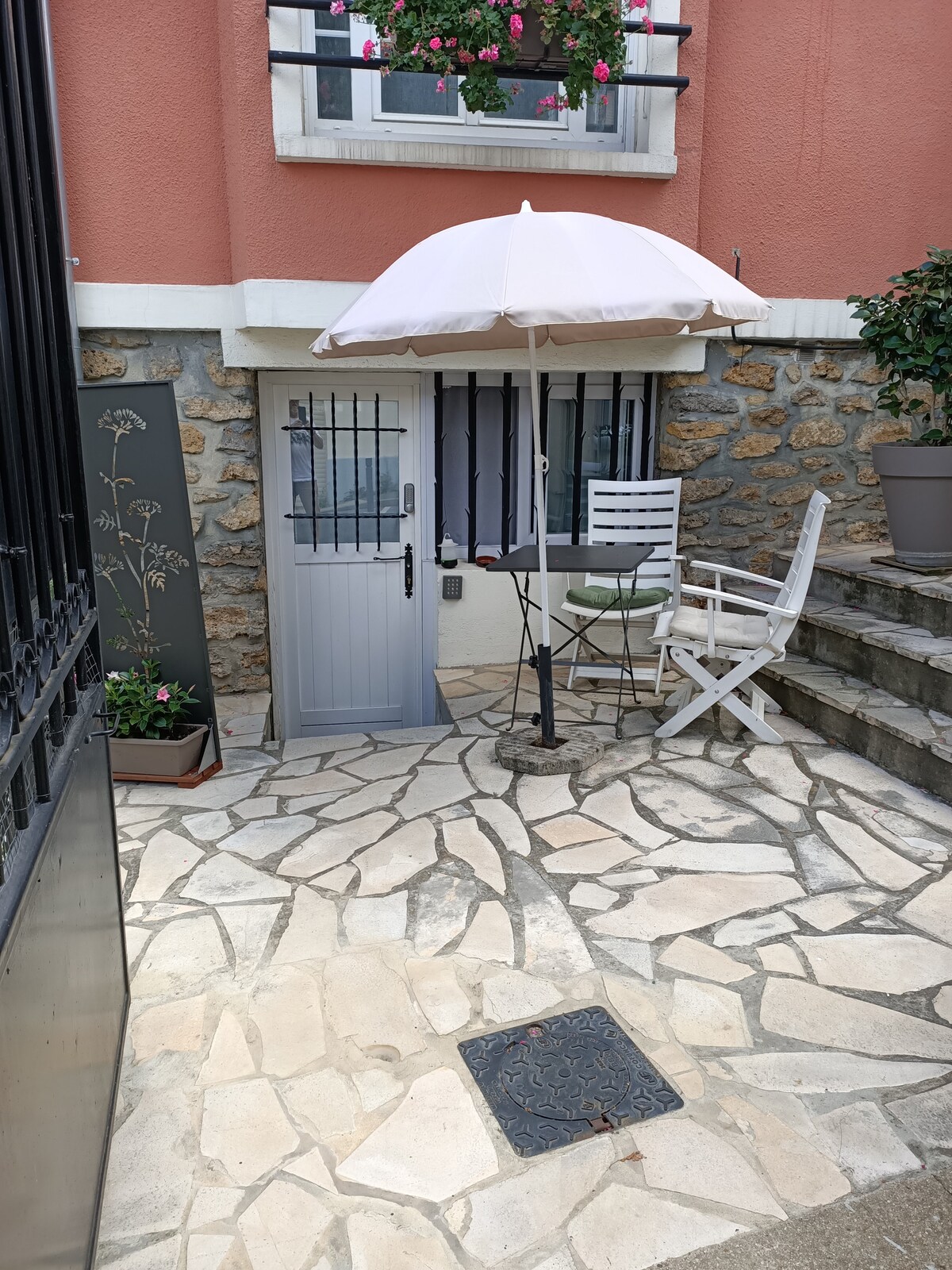 A small outdoor area is shown, featuring a stone-paved surface and a table with an umbrella. A white chair is positioned nearby. The entrance to the studio is visible, framed by a flowering plant and decorative elements against the warm-colored wall.