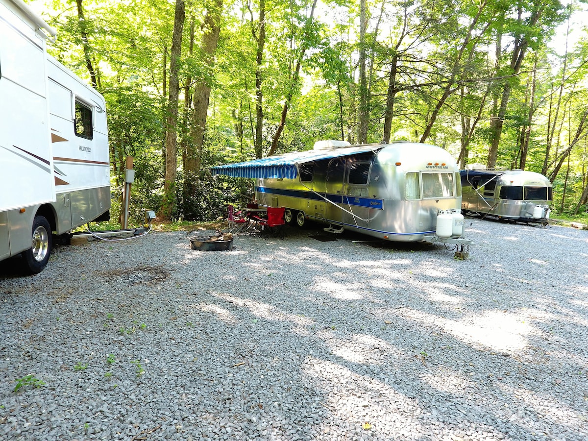 A vintage Airstream is parked on a gravel surface, surrounded by lush trees. The shiny exterior reflects sunlight, while a covered awning provides shade. Nearby, another Airstream and an RV are visible, contributing to a rustic camping atmosphere.