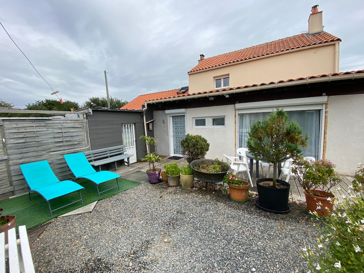 An outdoor terrace area is shown, featuring two turquoise lounge chairs positioned on gravel, surrounded by potted plants of various sizes. A gray wooden table and benches are visible in the background, with a white door leading to the interior of the property.