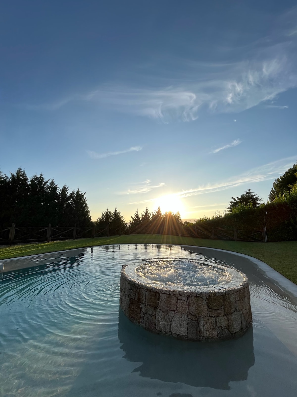 The image captures a tranquil swimming pool with a circular stone feature in the center. Gentle ripples are seen on the water's surface, reflecting the warm hues of the sunset. Lush greenery surrounds the pool, enhancing the serene outdoor setting.