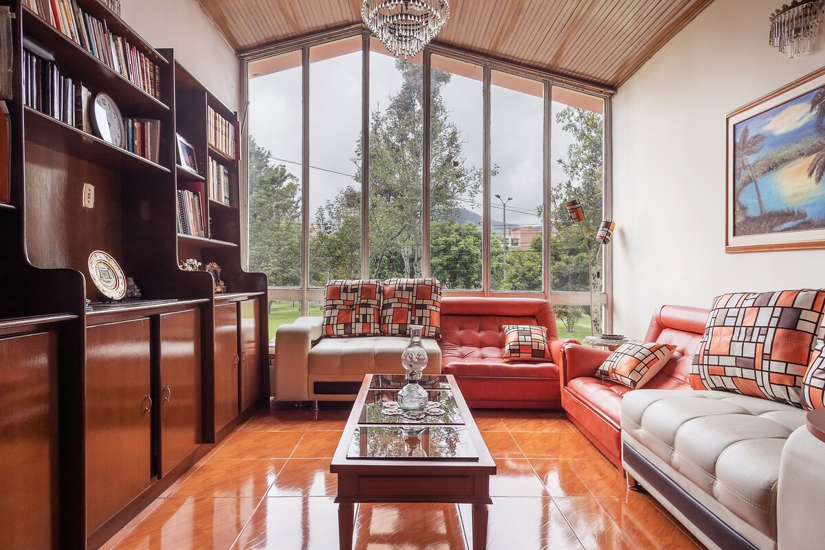 The living area features large windows that allow ample natural light to fill the space. A mix of red and beige seating options, accented with decorative cushions, surrounds a glass coffee table. A wooden bookshelf is positioned against the wall, displaying various books and decorative items.