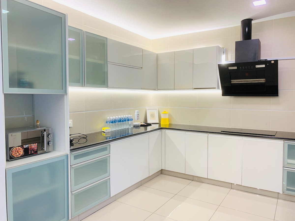 A modern kitchen is presented, featuring sleek white cabinetry and minimalist design. A black countertop contrasts with the glossy cabinets, and essential appliances including a microwave are visible. Bottled water is neatly arranged on the counter, and soft lighting enhances the spacious feel.