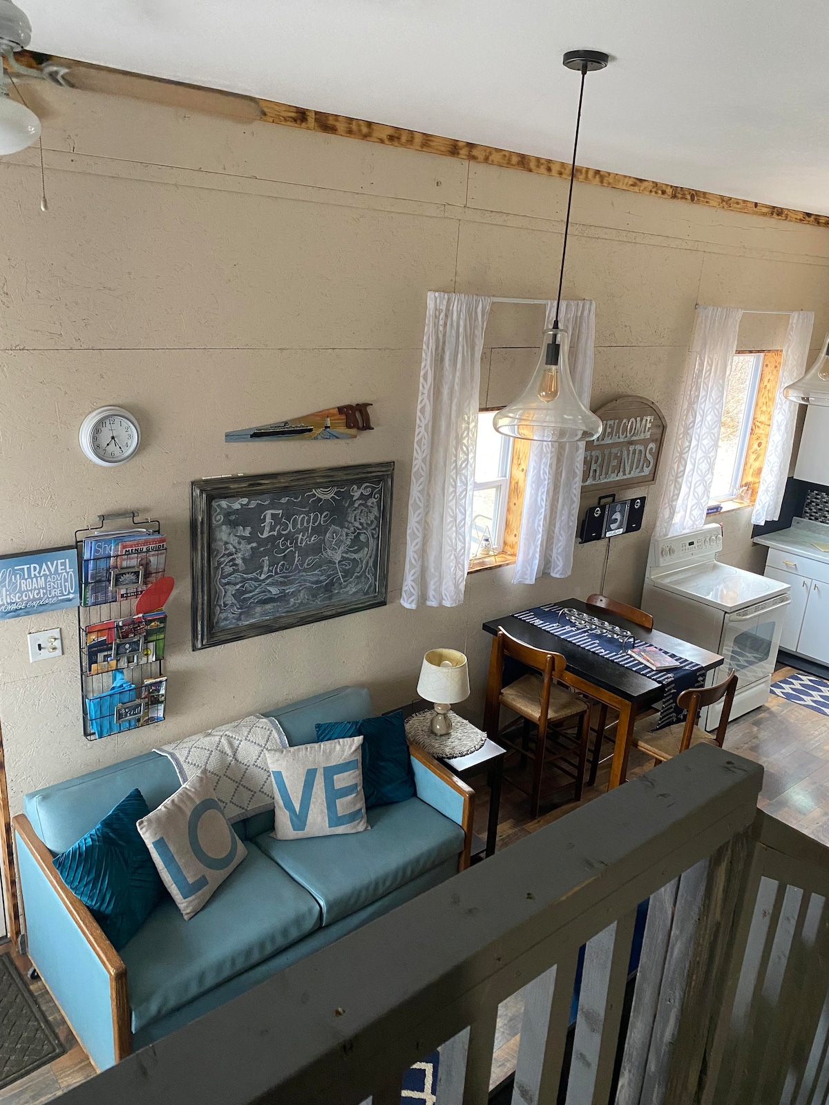 An inviting living area is highlighted by a blue couch adorned with decorative pillows. A wall-mounted chalkboard offers a welcoming message, while two windows provide natural light. A small dining area is positioned adjacent to the cozy space, featuring a table and chairs.