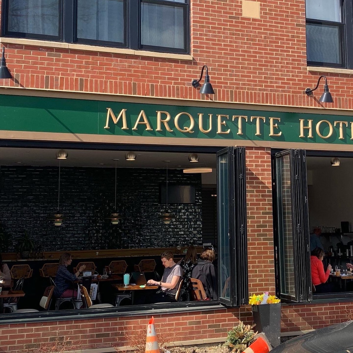 The exterior of the Marquette Hotel is prominently displayed, featuring a green awning with the hotel name. Large windows allow natural light into the cafe area, where guests are seated. A modern ambiance is suggested by the pendant lights and decorative interior visible through the windows.