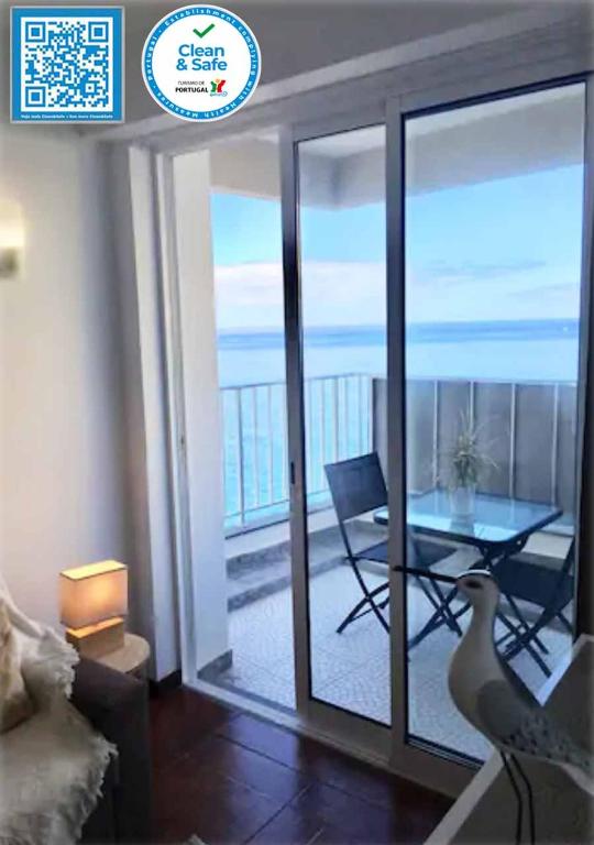 A bright living space features sliding glass doors that open to a small balcony with sea views. A cozy seating area includes a lamp and decorative accents. The balcony holds a table and two chairs, allowing for relaxation while enjoying the ocean breeze.