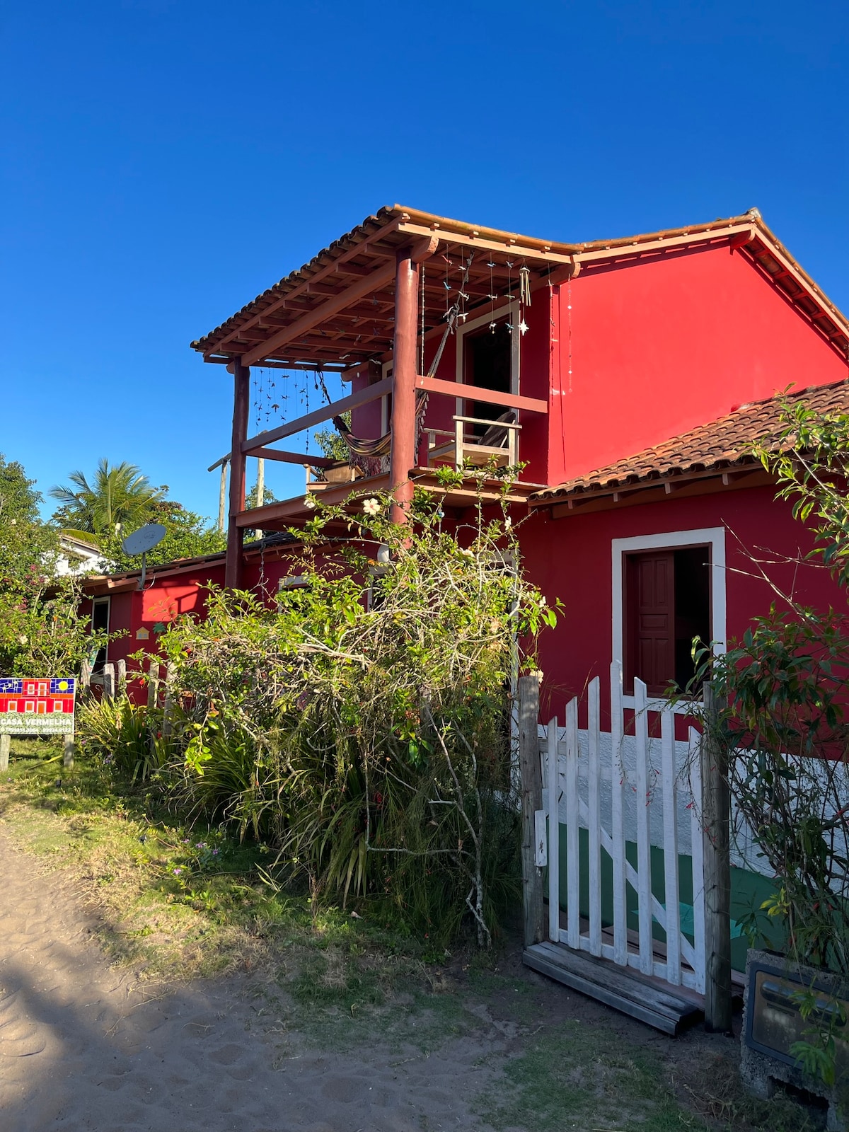 A vibrant red two-story house is depicted with a welcoming porch featuring wooden posts and a decorative railing. Lush greenery surrounds the house, enhancing its inviting exterior, while a clear blue sky provides a bright backdrop. A pathway leads up to a white fence.