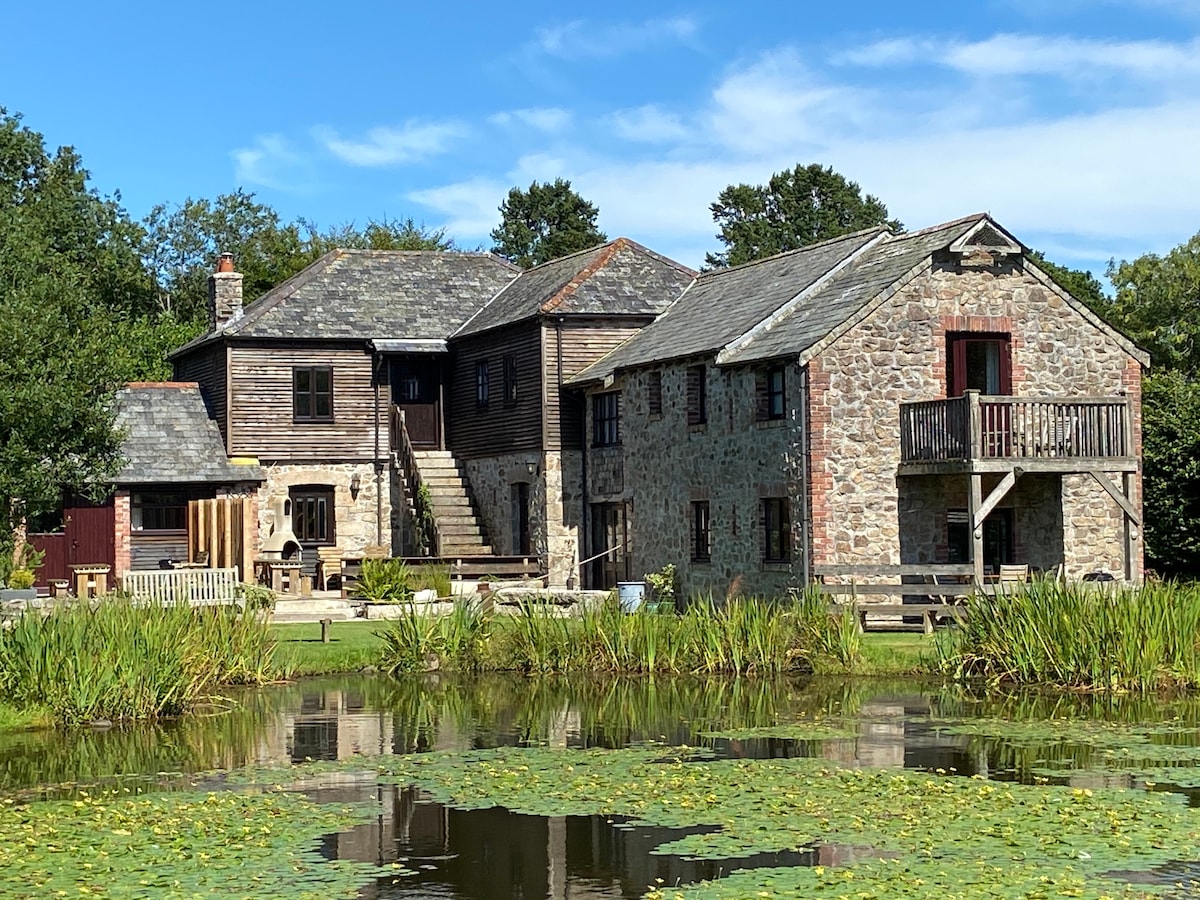 The exterior of Gulliver Cottage is showcased against a blue sky, with a blend of stone and wooden structures. Lush green surroundings are complemented by a tranquil pond dotted with lily pads, providing a serene backdrop for the cottage's multiple levels and outdoor spaces.