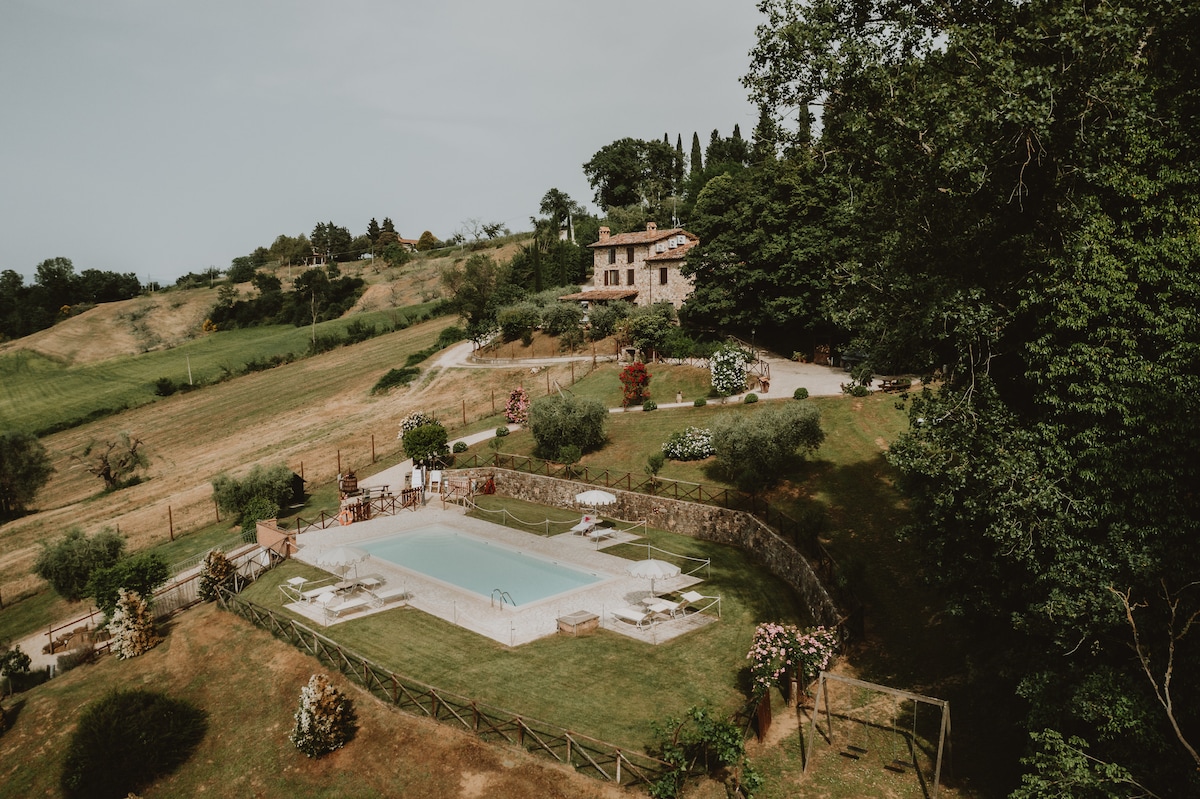 A spacious outdoor area features a well-maintained swimming pool surrounded by landscaped gardens and lush greenery. The villa is visible in the background, set against a scenic hillside that showcases rolling fields and trees.