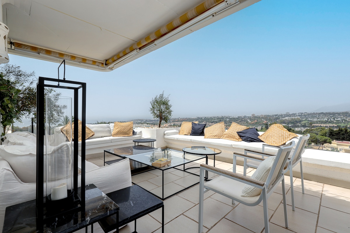 A spacious terrace features comfortable seating with decorative cushions, offering views of the surrounding landscape. A glass coffee table complements the seating area, while a lantern adds a subtle touch of ambiance. The bright sky provides a serene backdrop to the outdoor space.