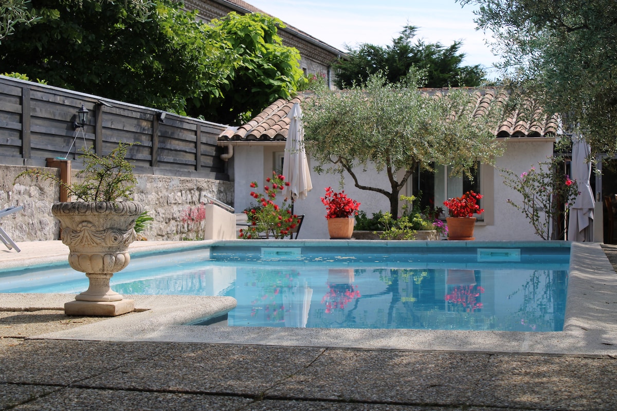 A private pool is surrounded by a paved terrace, featuring blooming flowers in pots and an olive tree. Umbrellas provide shade, and the charming exterior of a nearby building is visible in the background, creating a serene outdoor space.