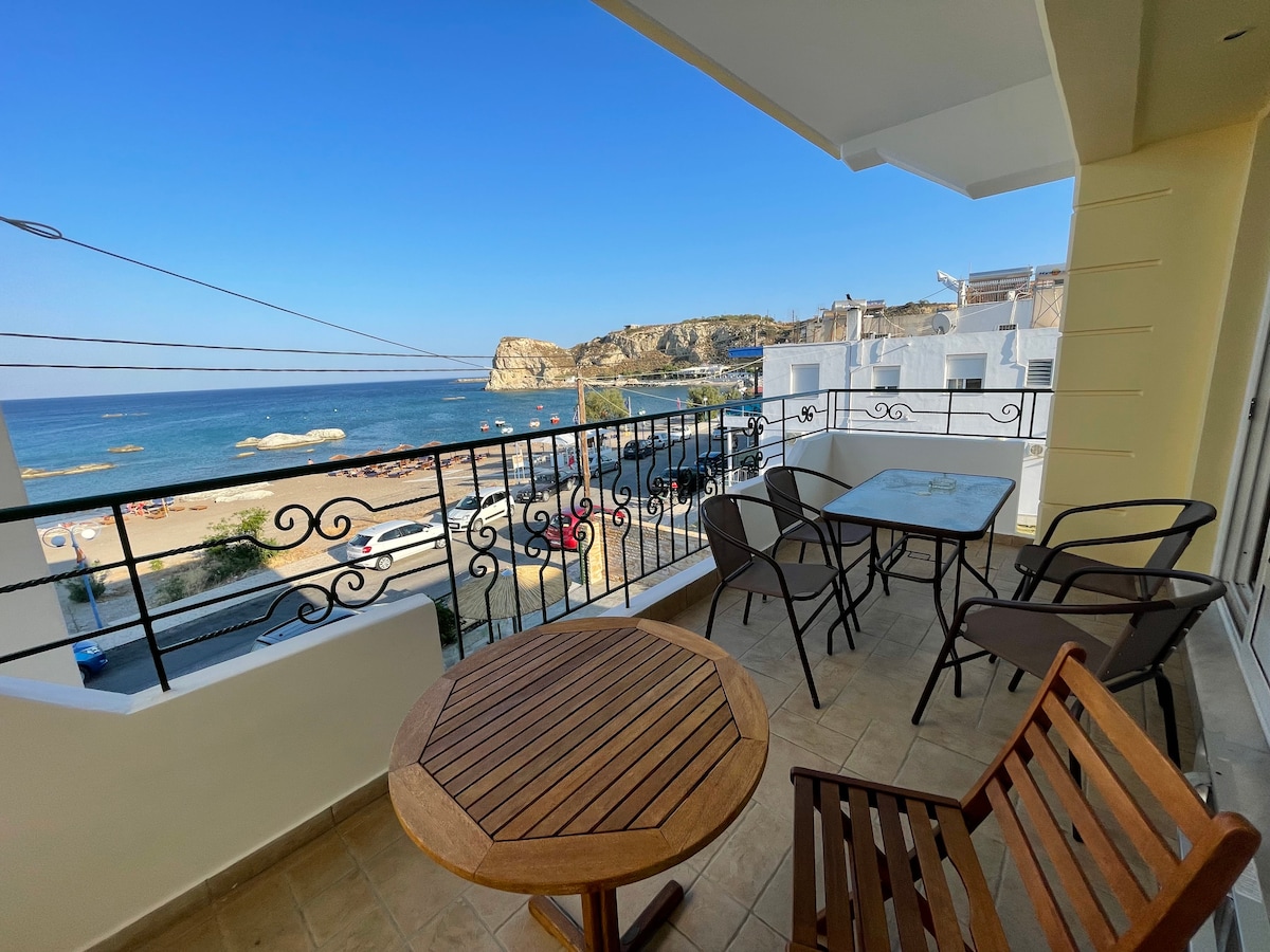 A spacious balcony offers a direct view of the seafront, featuring a wooden table surrounded by four chairs. The calm sea and distant cliffs are visible in the background, framed by clear blue skies. The balcony's railing adds to the scenic outlook.