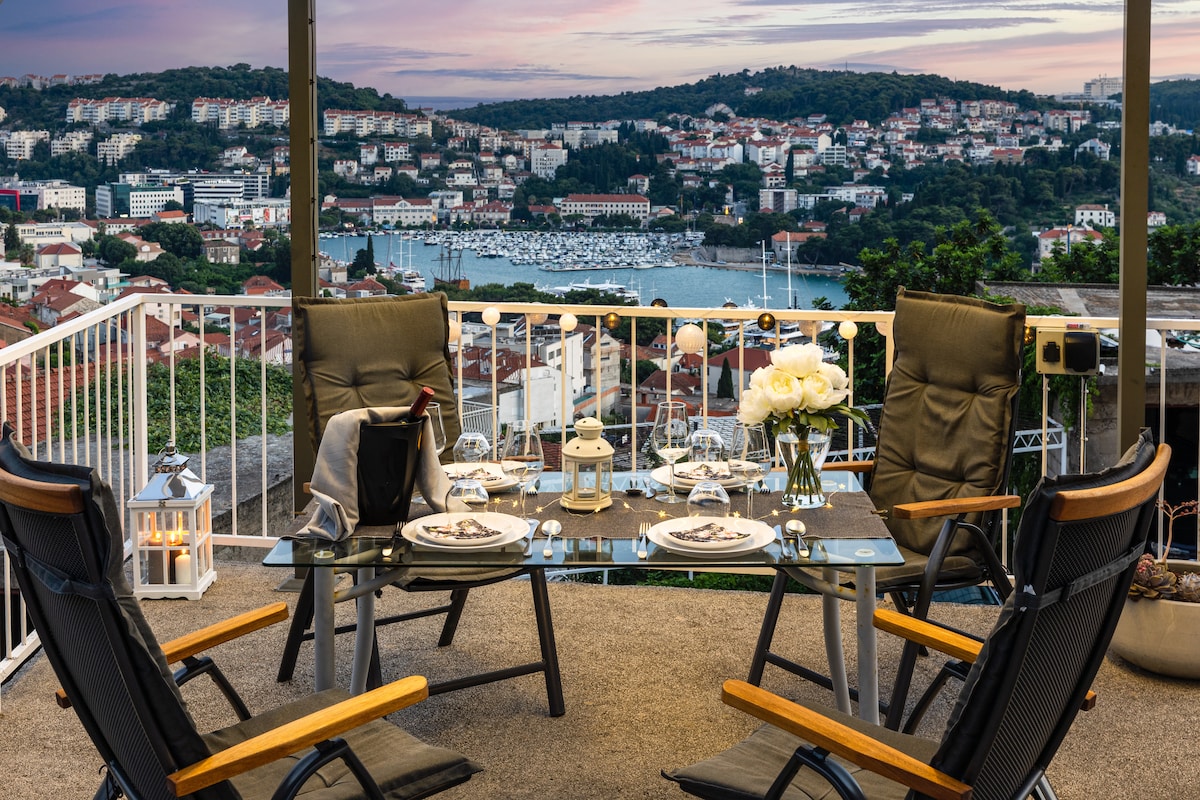 A dining space is presented on a terrace with a glass table set for a meal, surrounded by four reclining chairs. A vase of white flowers and lanterns add a serene touch. The view includes the Gruž bay and surrounding hills, enhancing the outdoor experience.