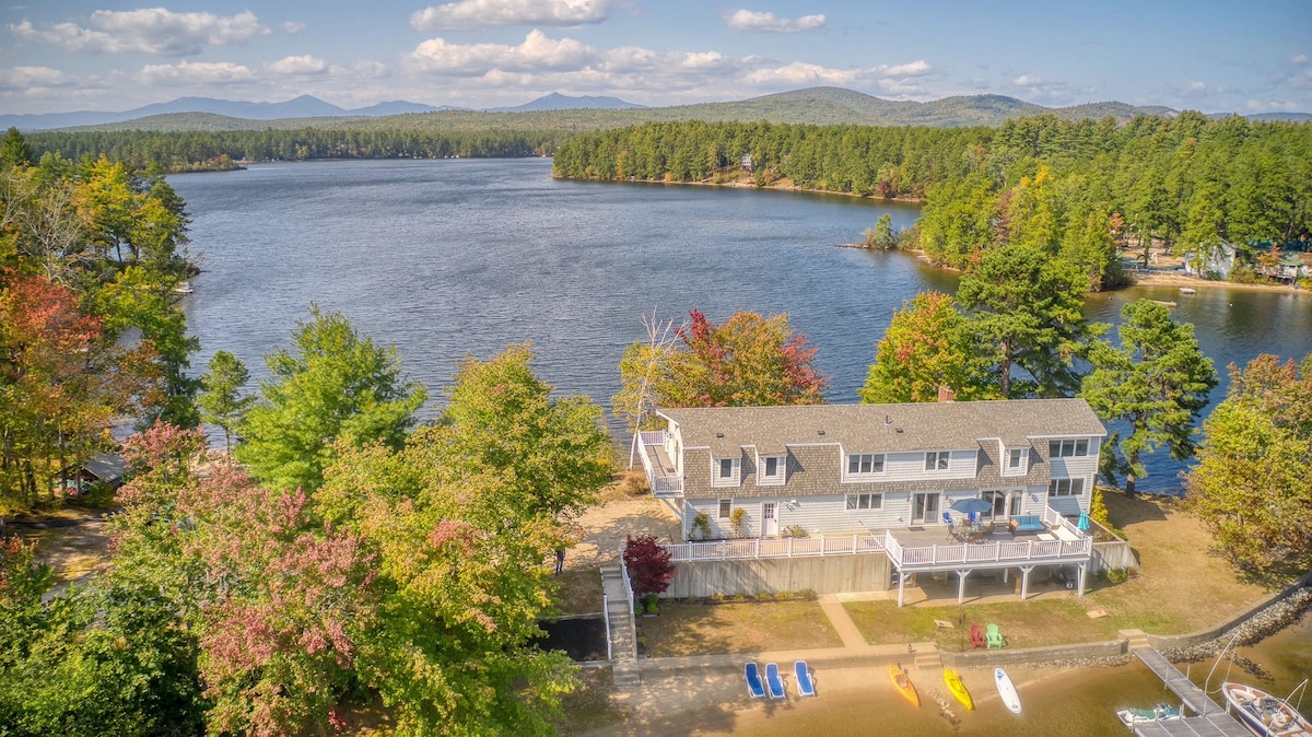 A waterfront view captures the spacious home set against a backdrop of trees and distant mountains. The property features a private sandy beach area, complemented by a variety of watercraft, including kayaks and paddle boards, arranged along the shore.