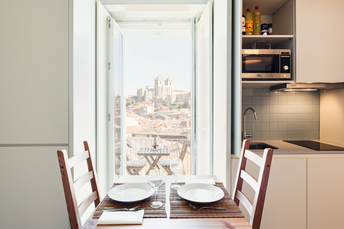 An open kitchen area showcases modern cabinetry and appliances, including a microwave and sink. A dining table is set for two with plates and utensils. Natural light floods in from the double doors that lead to a balcony, offering views of the city skyline.