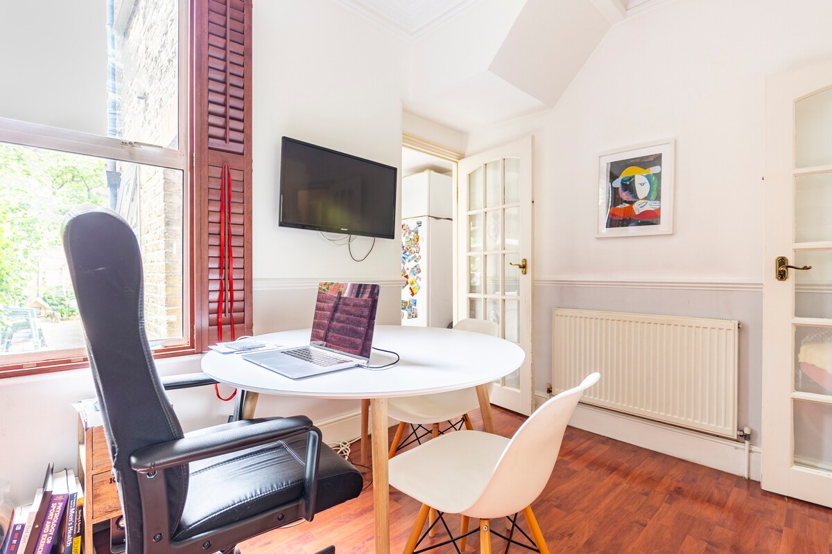 A bright workspace features a circular table with a sleek laptop. A comfortable chair is positioned next to the table, complemented by an additional chair. Natural light enters through a window, and a wall-mounted television is visible. Nearby, a colorful artwork adds a touch of creativity.