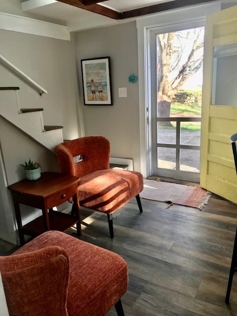 A welcoming entry area features two orange accent chairs and a small wooden table. Natural light pours in through a glass door leading to the outside, illuminating the gray walls. A staircase is visible in the background, while a framed artwork adds a personal touch.