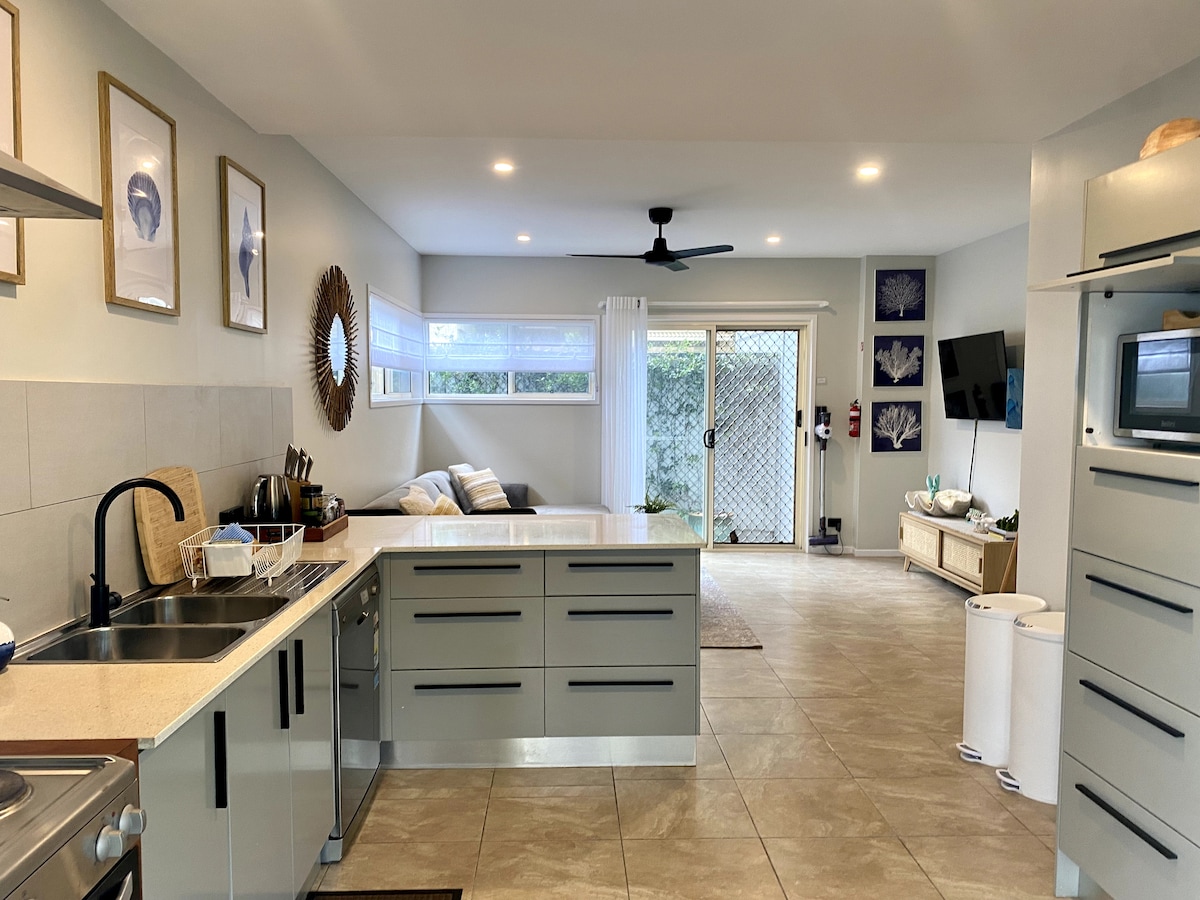 A modern kitchen is paired with an open living area, showcasing sleek cabinetry and a spacious island. Natural light enters through large windows, illuminating the room. A comfortable seating area and doors lead to an outdoor space, creating a seamless transition.