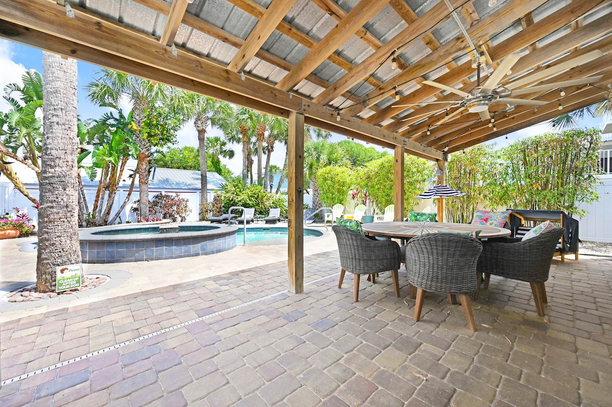 An outdoor patio area features a large dining table surrounded by four woven chairs. A ceiling fan hangs above, providing airflow. Palm trees and lush greenery surround a sparkling pool, highlighting the inviting outdoor atmosphere. Paved stones create a warm and grounding surface.
