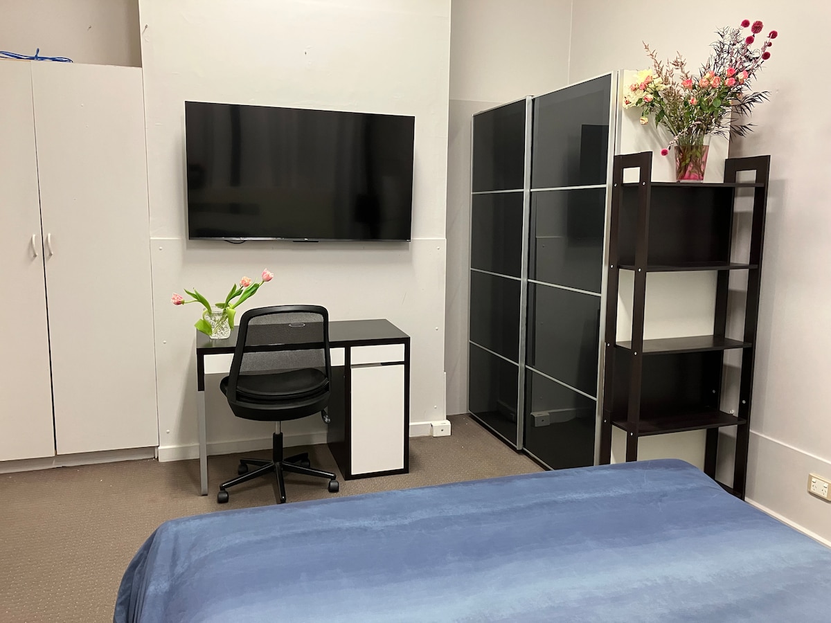 A spacious room includes a queen size bed with a blue coverlet, a large wall-mounted TV, and a work desk with a chair. Storage is provided by a sliding door wardrobe and an additional shelving unit. Fresh flowers add a touch of color to the space.