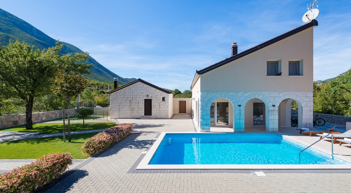 A modern villa is situated on a spacious estate, featuring a sparkling blue pool with a shallow entry. An additional stone structure is visible in the background. Lush greenery surrounds the area, complementing the serene mountain backdrop and providing a peaceful atmosphere.