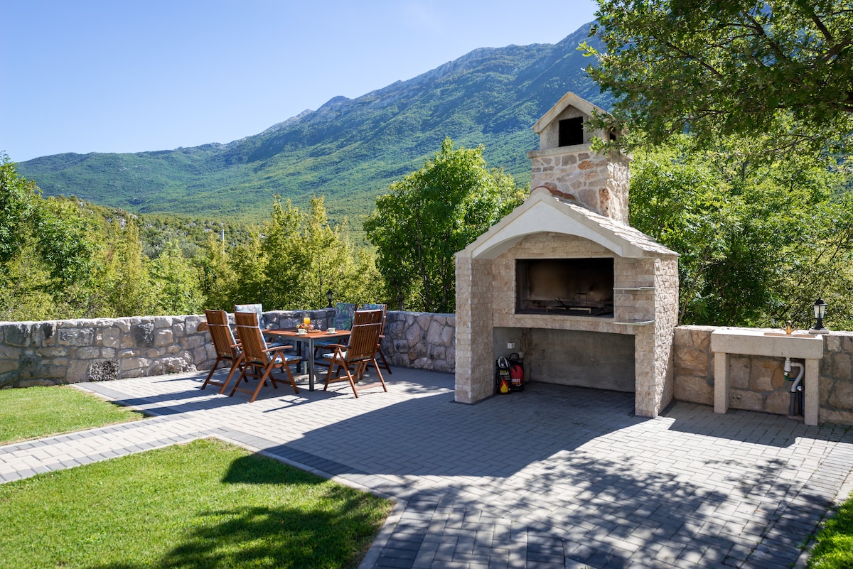 An outdoor barbecue area is situated on a paved patio, featuring a stone grill and a wooden table surrounded by six chairs. Lush greenery and a mountainous backdrop enhance the setting, providing a peaceful and natural environment for outdoor dining.