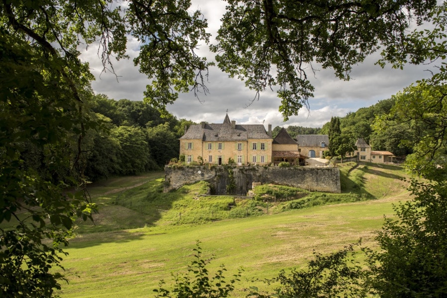A historic château is nestled among lush greenery, revealing stone walls and multiple gabled roofs. Surrounded by a sprawling estate, the structure's architectural details are framed by foliage, emphasizing its serene setting on an elevated terrain.