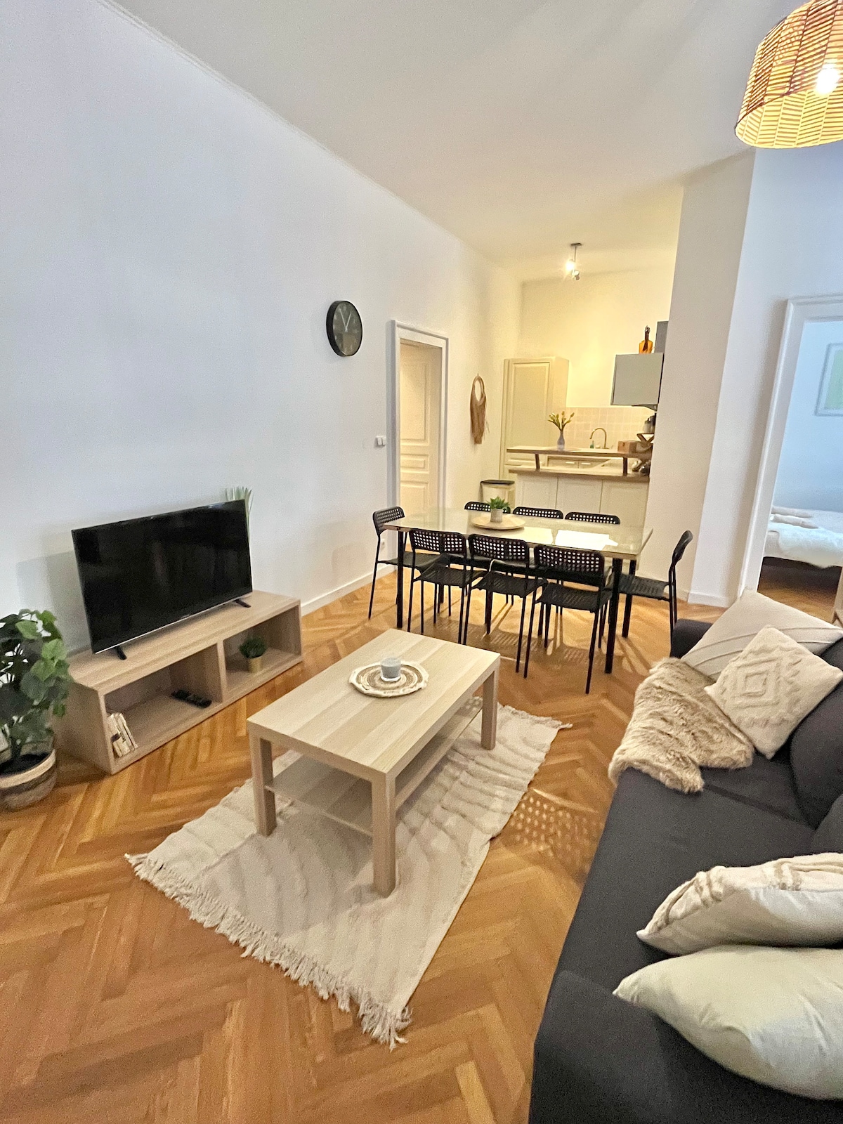 The living room is designed with a flat screen TV and a cozy seating area that includes a dark sofa adorned with cushions. A light wooden coffee table is centered on a light rug, while a dining table with six black chairs is visible in the background.