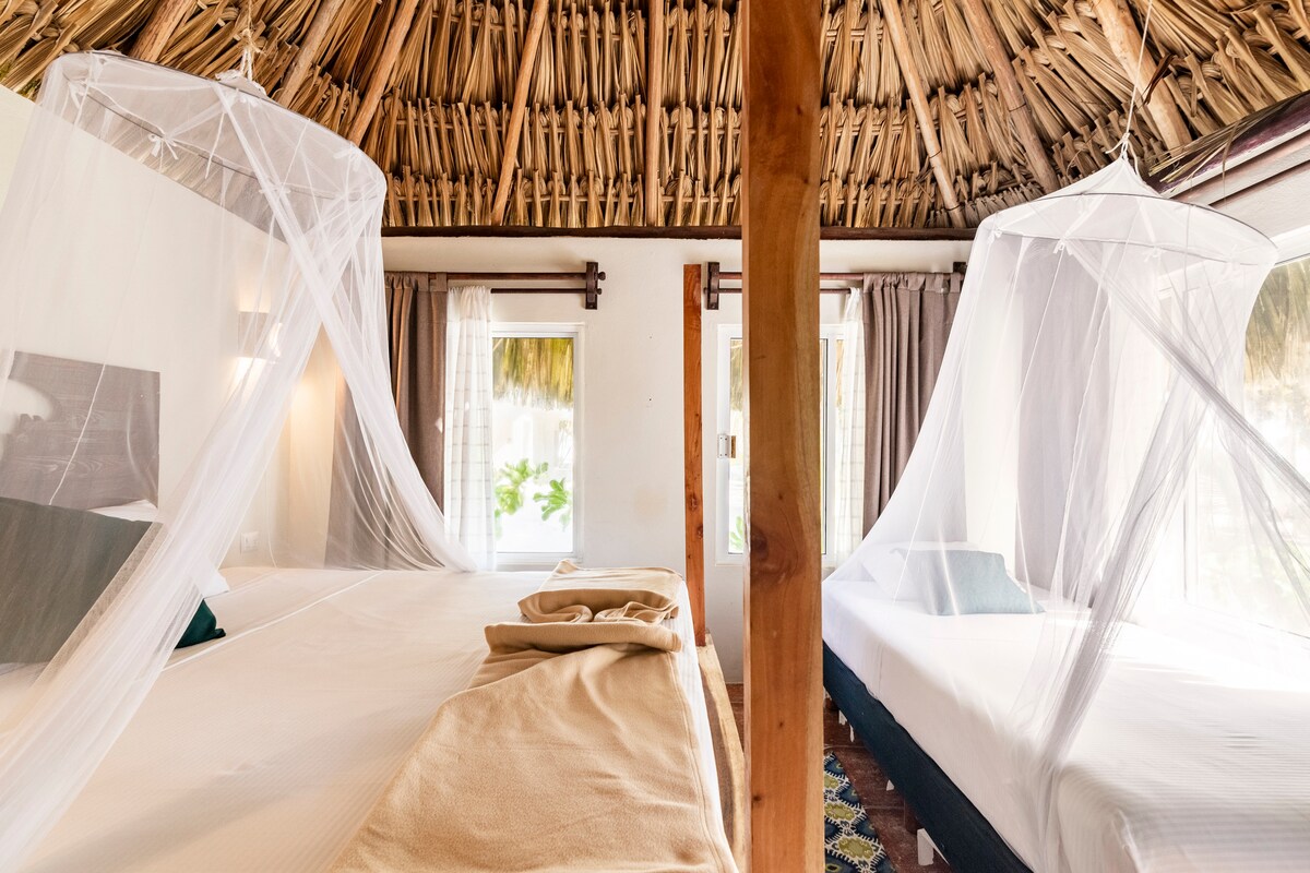 A rustic interior features two beds, each draped with mosquito nets. Natural light filters through large windows dressed in light curtains, illuminating the room. The thatched roof adds an organic touch, while the earthy tones of the bedding create a serene environment.