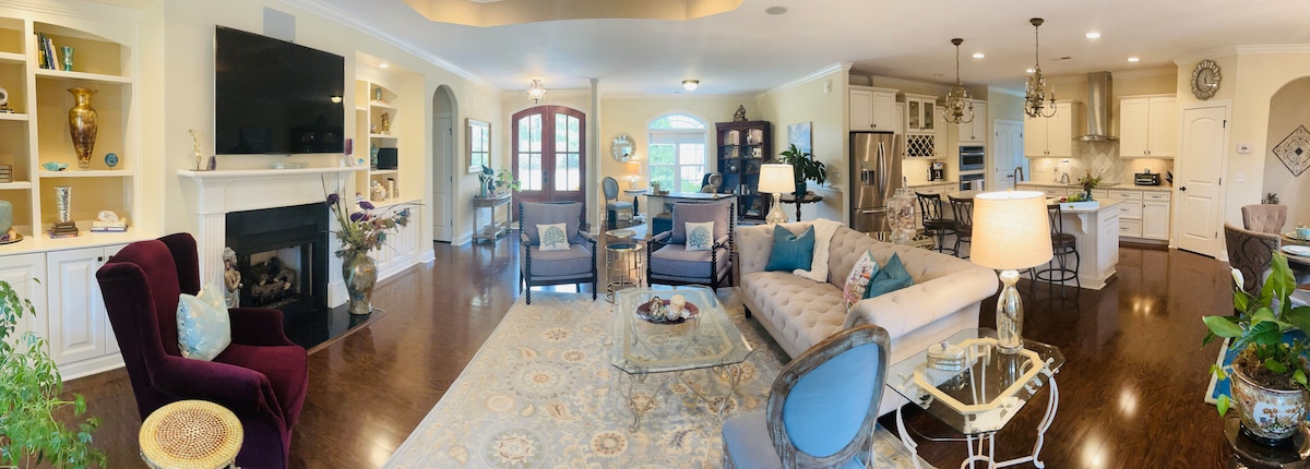 An open living area features a spacious layout with comfortable seating arrangements, including a plush sofa and elegant armchairs. A large TV is mounted above a cozy fireplace. Light floods in through multiple windows, accentuating the warm color palette and tasteful décor.