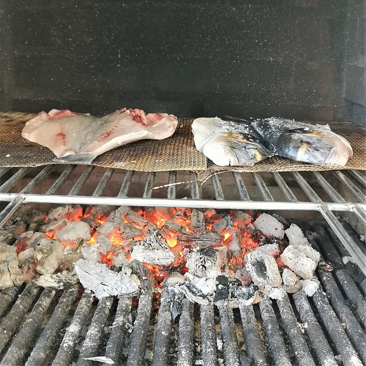 Fish fillets are being grilled over red-hot coals, resting on a woven mat to prevent sticking. The flames and glowing embers create a warm ambiance, indicating a cooking process that emphasizes traditional grilling methods.
