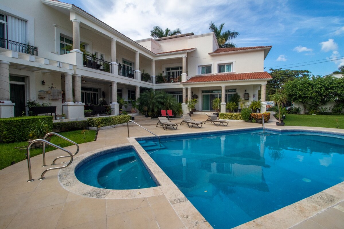 An outdoor area is presented featuring a large pool surrounded by loungers on a tiled deck. The building's two floors are visible, with multiple balconies framed by lush greenery. Palm trees and well-maintained grass enhance the inviting atmosphere of the space.