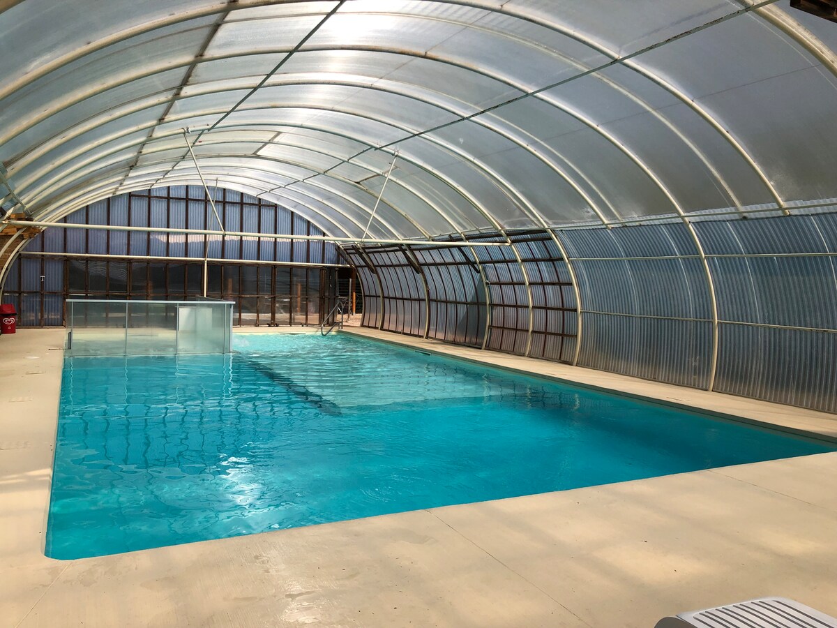 An indoor swimming pool is displayed under a translucent canopy, allowing natural light to illuminate the clear blue water. The pool area features a smooth, light-colored deck, creating an inviting space for relaxation. Surrounding elements suggest a well-maintained setting.