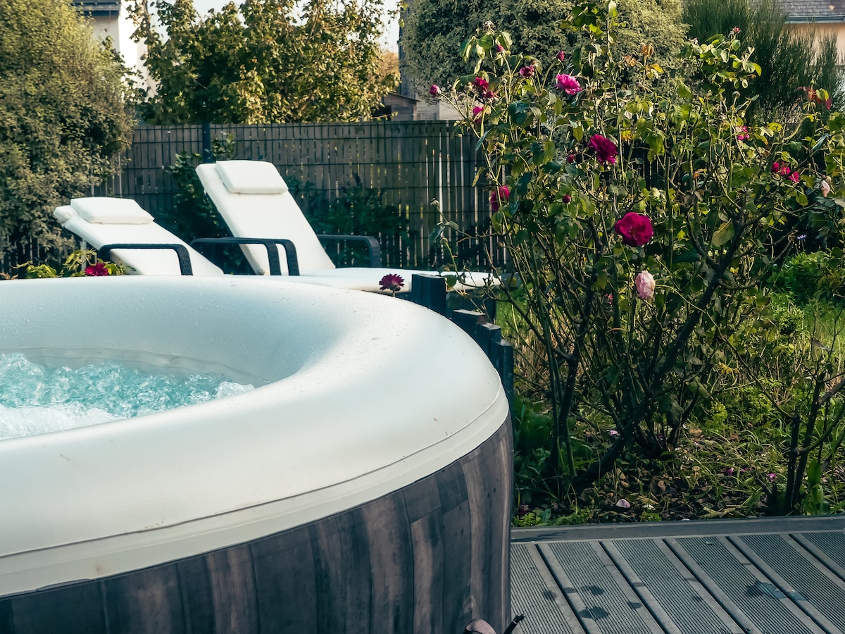 An outdoor spa is positioned next to blooming roses in a lush garden. Two white lounge chairs are visible nearby, offering a space for relaxation amidst greenery. The spa's surface creates gentle ripples, reflecting the surrounding nature.