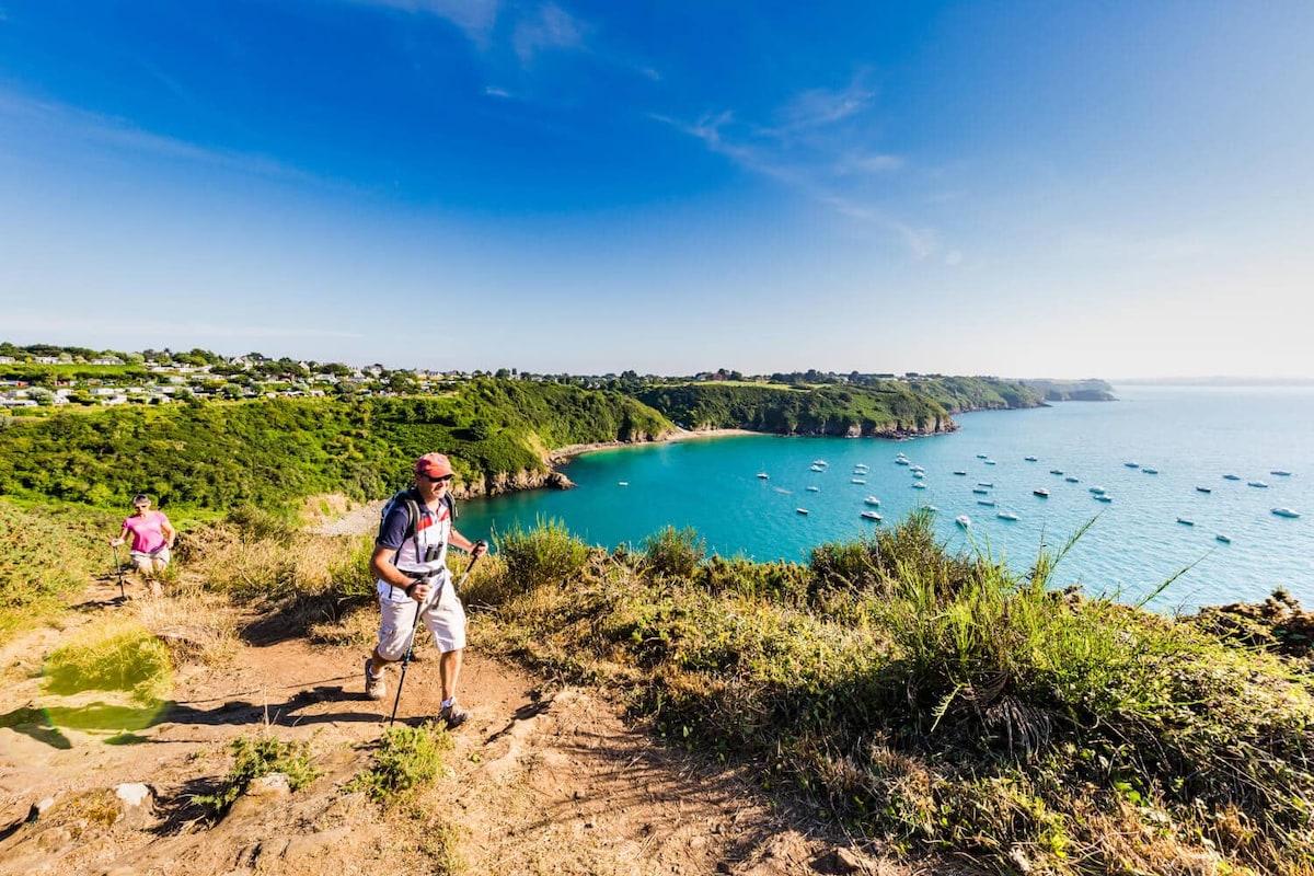 A scenic coastal landscape features a hiker navigating a trail overlooking a tranquil blue bay dotted with boats. Lush green hills frame the picturesque shoreline under a clear, sunny sky, enhancing the natural beauty of the area.