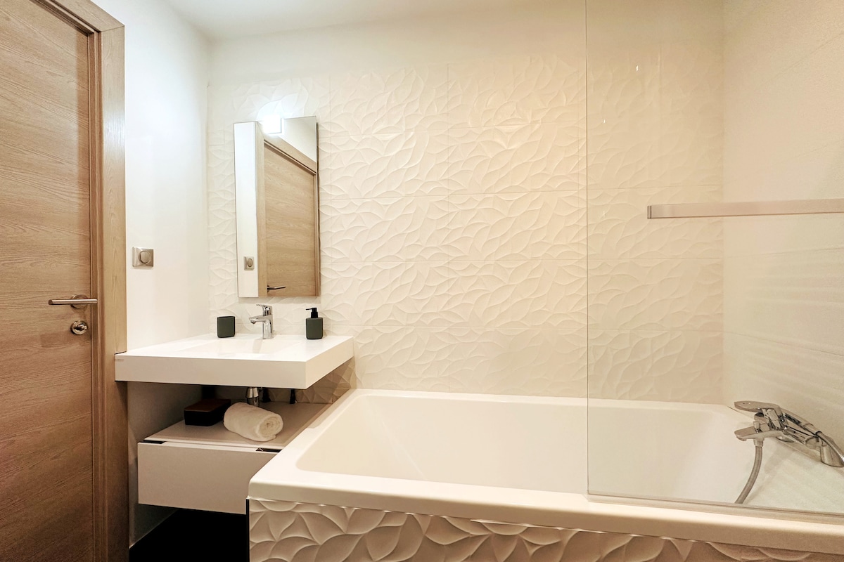 The bathroom features a clean, contemporary design with a full-length bathtub and a glass shower enclosure. A modern vanity with a mirror and sink is complemented by shelves below. Light-colored walls with a textured pattern enhance the space, with subtle lighting provided overhead.