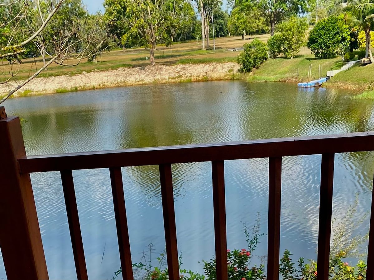 A serene lake view is visible from the balcony, framed by a wooden railing. The calm water reflects the surrounding greenery, including trees and shrubs. A small boat dock is seen in the distance, adding to the tranquil atmosphere of the outdoor setting.