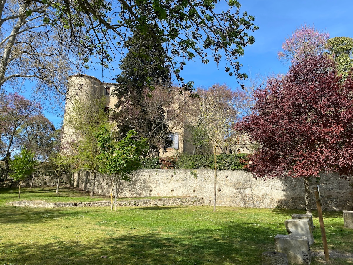 A scenic view of a stone structure is framed by lush greenery and trees in various shades, including a vibrant red one. The building features multiple windows and is set against a clear blue sky, creating a peaceful outdoor environment.