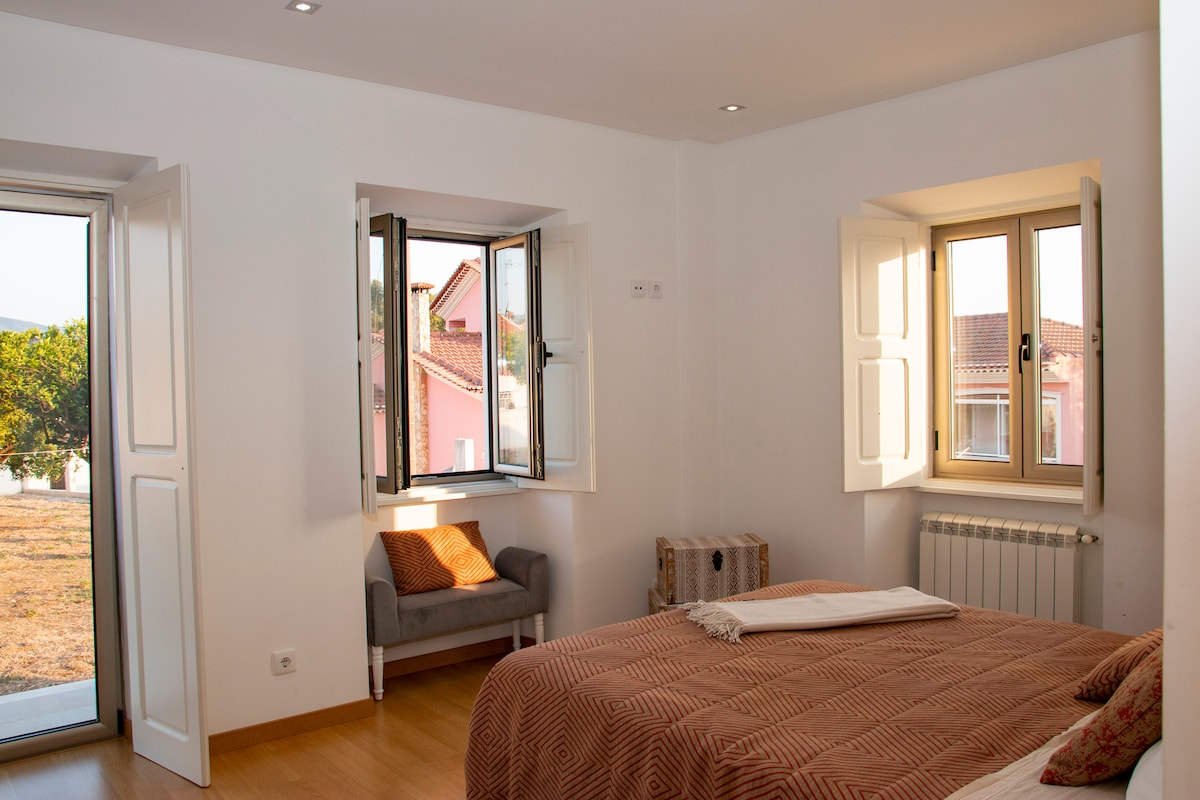 A bright bedroom is presented with two opening windows, enhancing natural light. A spacious bed is complemented by a cozy chair in the corner. A view of the village is visible through the windows, and wooden flooring contributes to the room's inviting atmosphere.