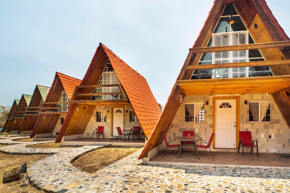 A series of triangular-shaped cabins feature wooden exteriors with red roofs. Each cabin has a front porch with chairs, and stone pathways lead between the units. The structure's design emphasizes a connection to the surrounding nature.