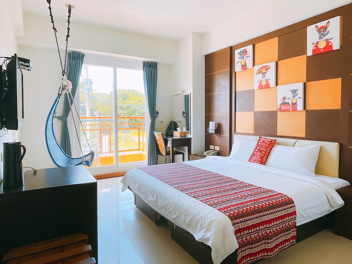 A well-lit room features a spacious bed with a patterned throw. A hanging chair is positioned near a large window that offers outdoor views. A desk and chair accompany a small workstation, complemented by artistic wall decorations and privacy curtains.