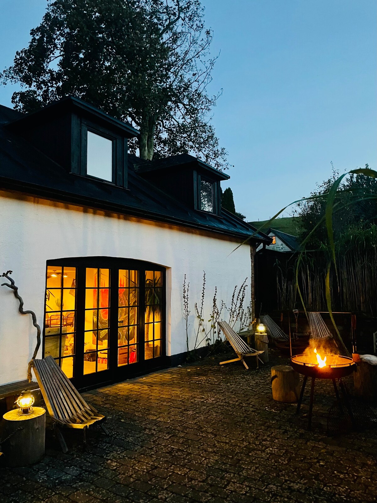 An inviting outdoor space is revealed at dusk, featuring a cozy fire pit surrounded by comfortable wooden lounge chairs. The white building with large glass doors reflects warm interior lighting, enhancing the relaxed atmosphere of the courtyard, which is framed by trees and low grass.